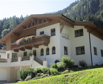 A traditional Austrian house with a large balcony and flower boxes. Surrounded by green hills and forests.