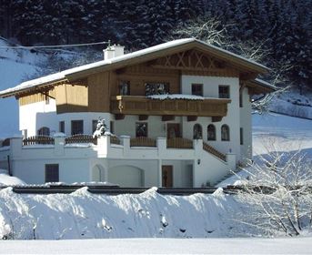 Ein großes, charmantes Holzhaus im Winter, umgeben von Schnee. Die Architektur hat alpine Elemente und es gibt Balkone und Fenster mit Blick auf die Umgebung.