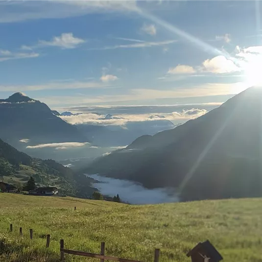 Eine malerische Berglandschaft mit sanften Hügeln und einem strahlenden Sonnenaufgang. Nebel schwebt über dem Tal und die Wolken sind teilweise am Himmel sichtbar.