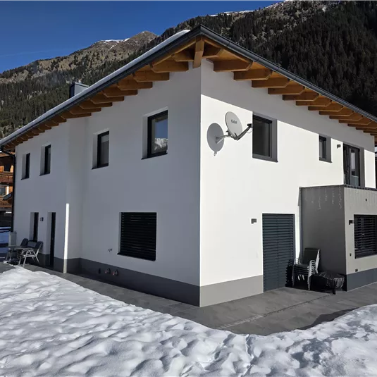Ein modernes, weißes Haus mit Holzdetails steht in einer verschneiten Umgebung. Im Hintergrund sind Berge zu sehen, die eine alpine Landschaft schaffen.