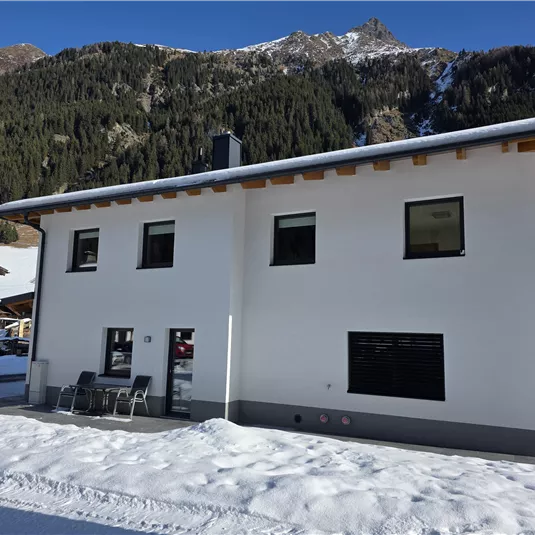 Ein modernes Haus in den Bergen, umgeben von Schnee. Die klare Himmel und die majestätischen Berge im Hintergrund ergänzen die alpine Landschaft.