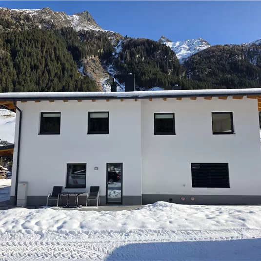 Ein modernes, weißes Haus steht in einer verschneiten Landschaft. Im Hintergrund sind Berge mit Wälder zu sehen.