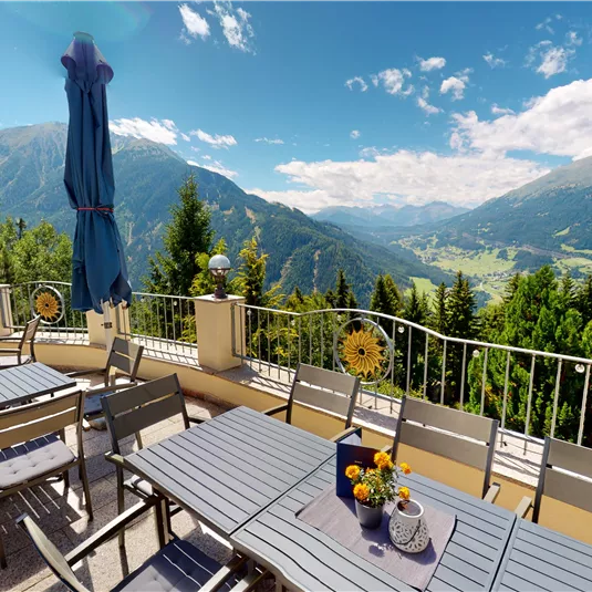 Eine Terrasse mit Tischen und Stühlen bietet einen herrlichen Blick auf die Berge und das Tal. Der Himmel ist blau mit wenigen Wolken, und die Umgebung ist grün und einladend.
