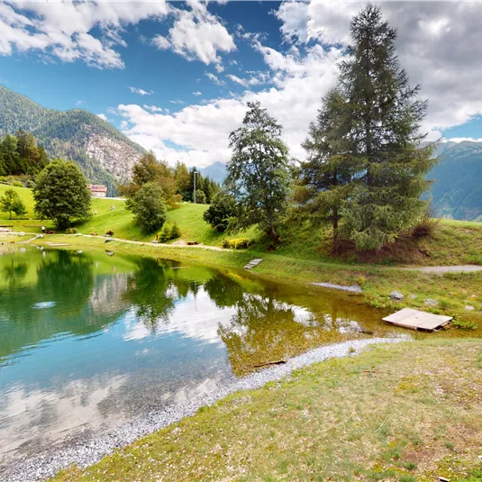 Ein ruhiger See umgeben von üppigem Grün und Bergen. Der Himmel ist teilweise bewölkt und reflektiert sich im Wasser.
