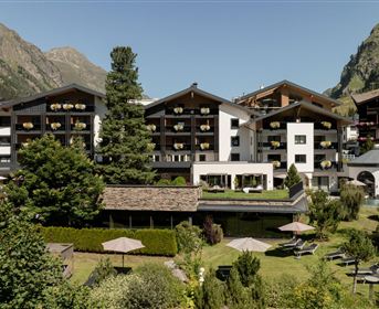 Ein modernes Hotelgebäude umgeben von grüner Landschaft und Bergen. Entspannte Atmosphäre mit Gartenbereichen und Sonnenschirmen.