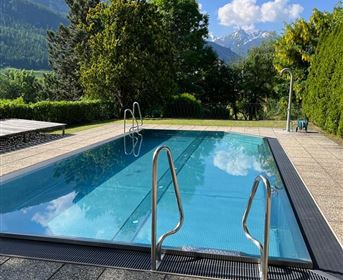 Ein klarer Swimmingpool in einer Gartenlandschaft mit Bäumen und Bergen im Hintergrund. Der Himmel ist blau und es ist sonnig.