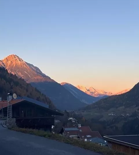 A picturesque mountain landscape at sunset with gentle hills and snow-capped peaks. In the foreground, there are some houses and a quiet road.