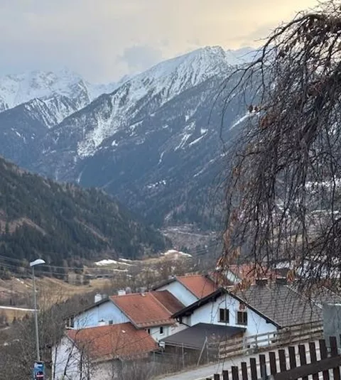 A picturesque mountain landscape with snow-capped peaks and rustic houses in the foreground. The sky is cloudy and the atmosphere is peaceful.