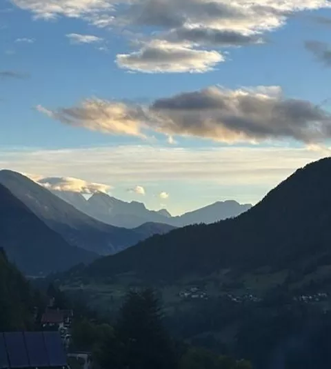 A picturesque mountain landscape with gentle hills and clouds in the sky. The sun is setting, bathing the scene in warm light.