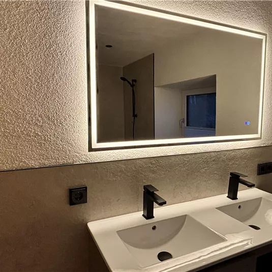 A modern bathroom with two sinks and a lit mirror. The walls are simply and elegantly designed.