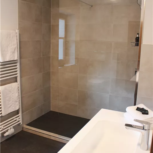 A modern bathroom with a walk-in shower and a sink. The walls are designed with neutral tiles and there are fresh towels.