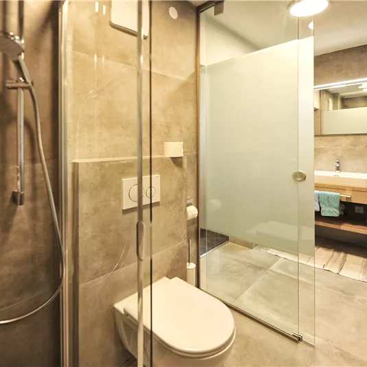 Ein modernes Badezimmer mit einer Dusche und einer klaren Glaswand. Die Wände sind in sanften Grautönen gehalten und es gibt Holzakzente.