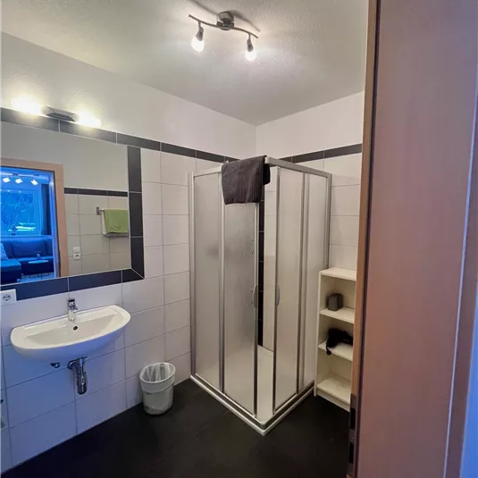 A modern bathroom with a shower, a sink, and a small shelf. The walls are brightly tiled and the floor is dark.