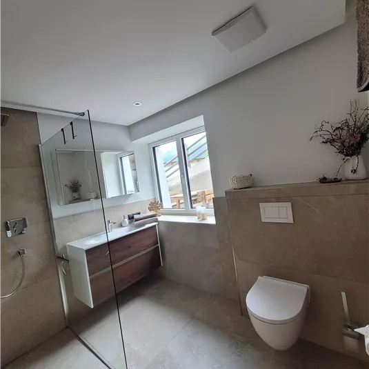 Ein modernes Badezimmer mit hellen Fliesen und einem großen Fenster. Es sind eine Dusche mit Glaswand, ein Waschbecken und eine Toilette zu sehen.