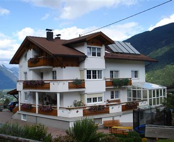 Ein modernes Mehrfamilienhaus mit einem Balkon und vielen Fenstern. Im Hintergrund sind schöne Berge und ein klarer Himmel zu sehen.