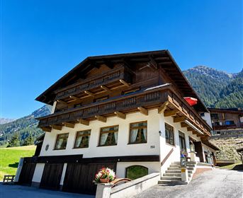A traditional chalet-style house with wooden-decorated balconies in a beautiful mountain landscape. The sky is clear and blue, surrounded by green meadows.