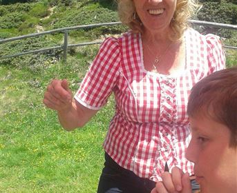 A smiling woman with curly hair is wearing a checkered shirt and is standing in a green environment. Next to her sits a child who looks thoughtful.