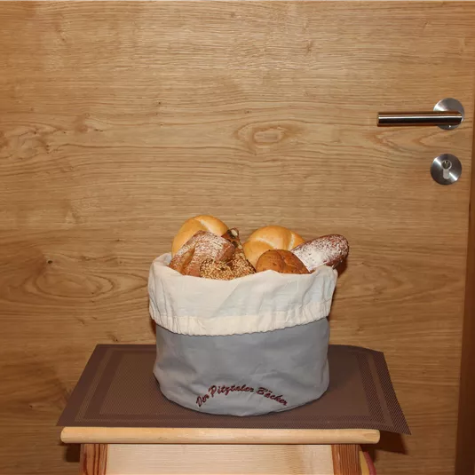 Ein Korb mit frischem Brot steht vor einer Holztür. Der Korb ist beige und grau, gefüllt mit verschiedenen Brötchen.