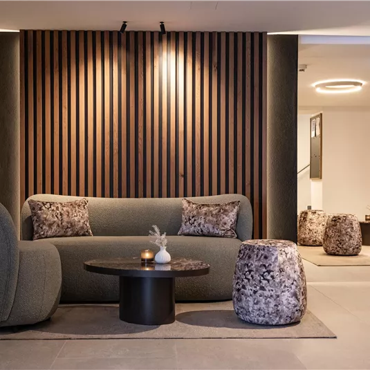 A modern living room with a cozy sofa and stylish armchairs. The walls are decorated with wood paneling, and soft lighting creates a relaxed atmosphere.