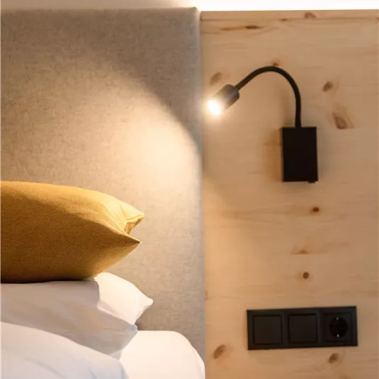 Ein modernes Bett mit einer gemütlichen Kopfstütze und einem schlichten Leselicht. Die Wand ist aus hellem Holz mit Steckdosen in der Nähe.