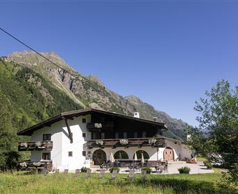 Ein rustikales Gebäude inmitten einer malerischen Berglandschaft. Die Sonne scheint und umgibt die Umgebung mit grünem Gras und Bäumen.