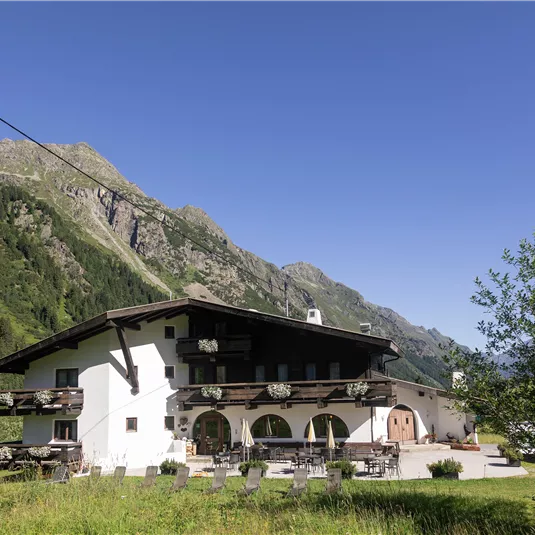 Ein rustikales Gebäude inmitten einer malerischen Berglandschaft. Die Sonne scheint und umgibt die Umgebung mit grünem Gras und Bäumen.