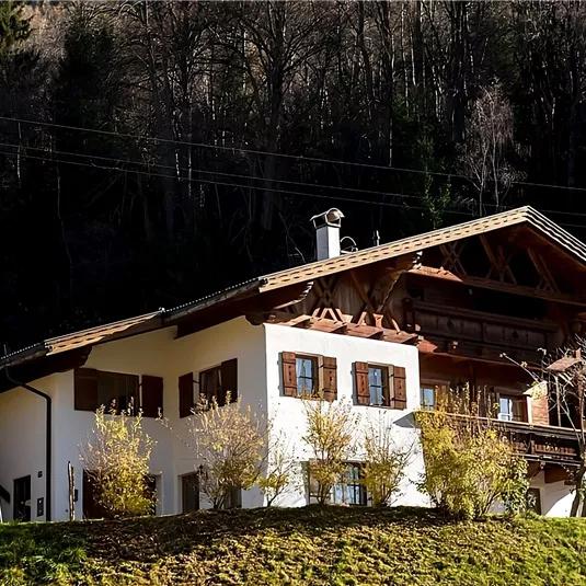 A traditional alpine house surrounded by trees and colorful autumn foliage. The architecture features a typical wooden gable and large windows.