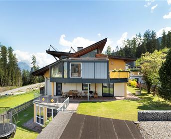 A modern house in a picturesque setting with mountains in the background. The garden is well-kept and offers space for outdoor activities.
