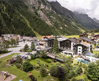 Eine malerische Berglandschaft mit mehreren Gebäuden und einem Garten. Im Hintergrund sind grüne Wälder und hohe Berge zu sehen.