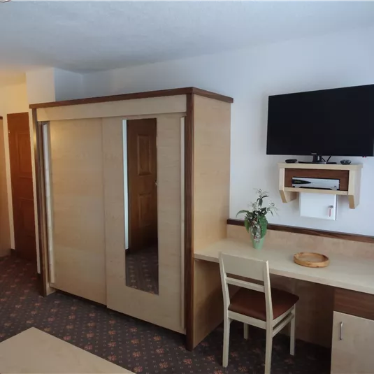 A cozy room with a large wardrobe, a desk, and a television on the wall. There is also a small table with a plant.