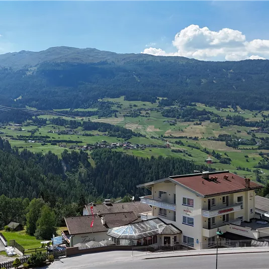 A beautiful view of green valleys and mountains under a clear blue sky. In the foreground, some buildings and roofs are visible.