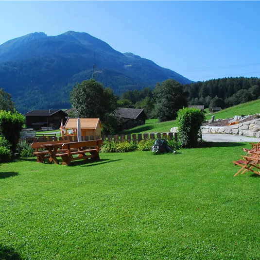Eine idyllische Wiese mit grünen Rasen und Aussicht auf Berge. Holzliegen und ein Tisch stehen im Freien, umgeben von Bäumen und Sträuchern.