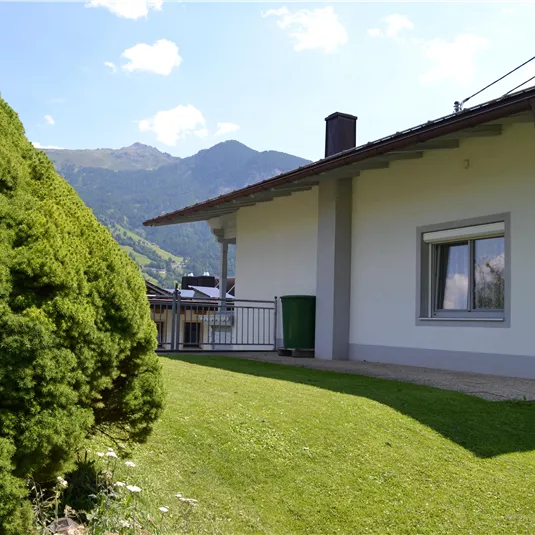 A modern house with green grass and a large tree in the foreground. In the background, mountains and a blue sky are visible.
