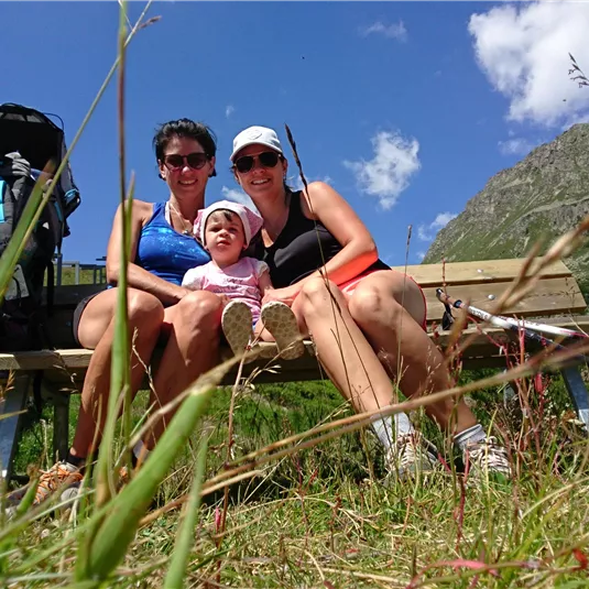 Three people are sitting on a bench in nature. In the background, mountains and a blue sky can be seen.