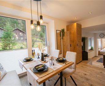 Ein modernes Esszimmer mit einem Tisch, der für vier Personen gedeckt ist. Große Fenster bieten Blick auf eine grüne Landschaft.