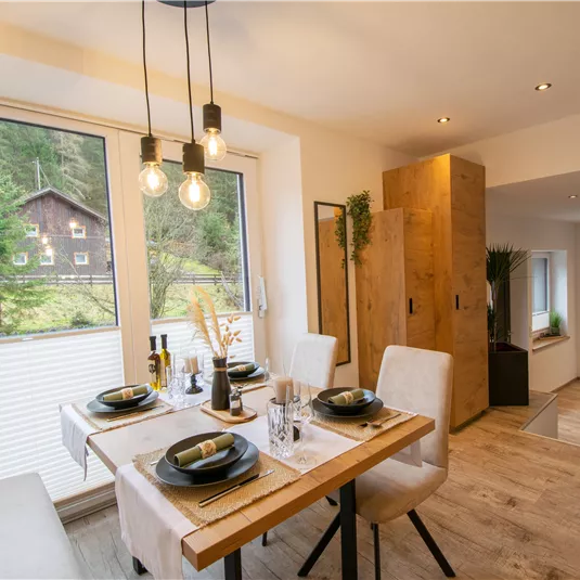 A modern dining room with a table set for four people. Large windows offer a view of a green landscape.