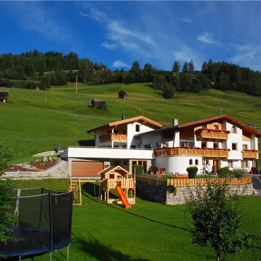 Ein schönes Haus in den Bergen mit einer großen grünen Wiese. Im Vordergrund sind ein Spielplatz und ein Trampolin zu sehen.