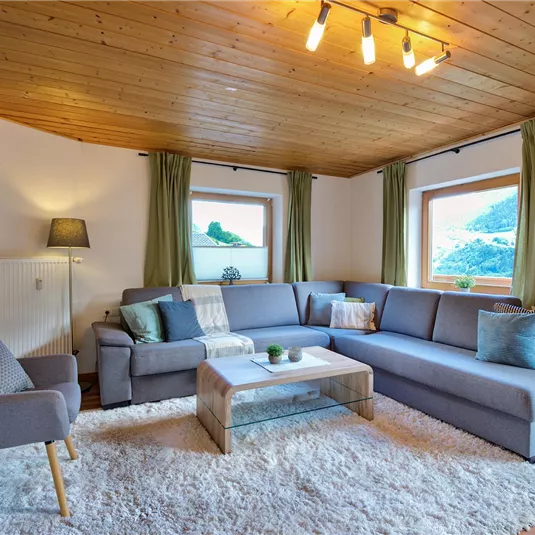 Ein modernes Wohnzimmer mit einer gemütlichen Couch und einem Couchtisch. Die großen Fenster lassen viel Licht herein und die Wände sind in sanften Farben gehalten.