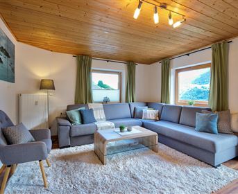 Ein modernes Wohnzimmer mit einer gemütlichen Couch und einem Couchtisch. Die großen Fenster lassen viel Licht herein und die Wände sind in sanften Farben gehalten.