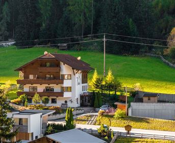 Ein charmantes Gebäude im alpinen Stil umgeben von grünen Wiesen und Bäumen. Im Hintergrund sind sanfte Hügel und ein klarer Himmel zu sehen.