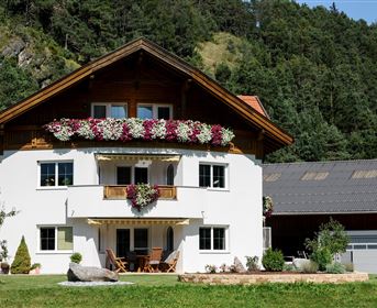 Ein schönes Haus mit blühenden Balkonblumen in einer grünen Landschaft. Im Hintergrund sind Bäume und eine Scheune zu sehen.