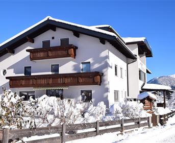 Ein schneebedecktes Haus mit Holzbalconys unter blauem Himmel. Umgeben von schneebedeckten Bäumen und einer ruhigen Winterlandschaft.
