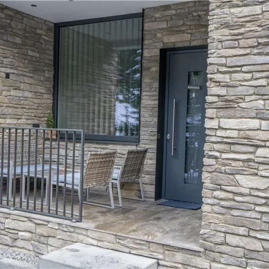 Entrance to a modern house with a stone facade. There is a small balcony with a table and chairs.