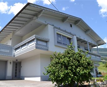 Ein modernes Haus mit einem großen Balkon und hellem Anstrich. Umgeben von Grün und einer schönen Landschaft.