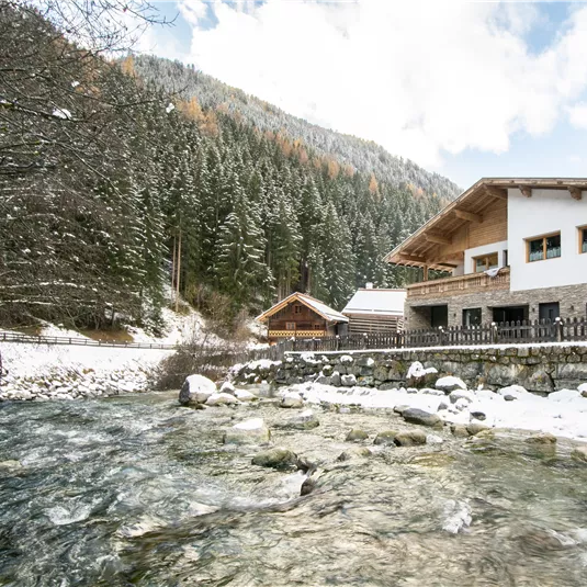 A snowy landscape with a clear river, surrounded by tall, green trees. In the background, a cozy house is visible.