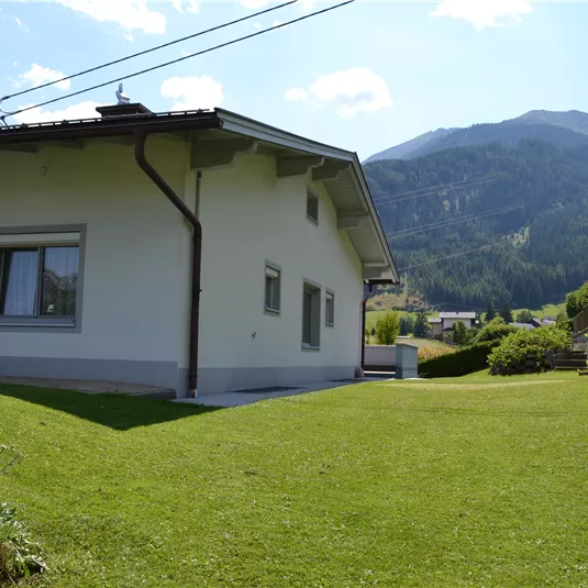 A modern house with a large garden and a stunning view of the mountains. The sky is clear, and the surroundings are green and inviting.