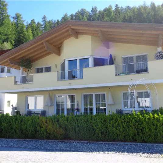 A modern house with a large balcony and a well-maintained hedge in the foreground. Surrounded by trees and a clear sky.