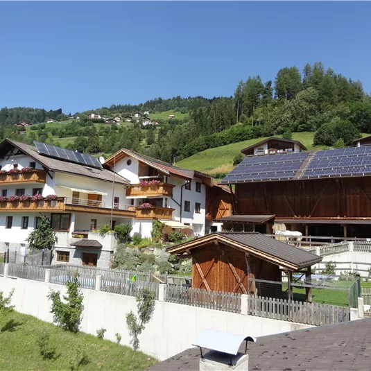 Eine malerische Berglandschaft mit traditionellen Holzhäusern und Solarpanelen. Grüne Wiesen und Bäume umgeben die Gebäude.