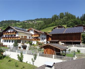 A picturesque mountain landscape with traditional wooden houses and solar panels. Green meadows and trees surround the buildings.