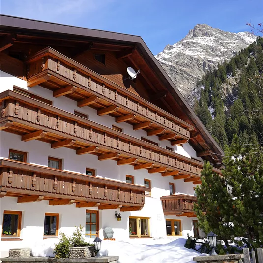 Ein traditionelles Alpenhaus mit Holzbalkonen steht vor einer schneebedeckten Landschaft. Im Hintergrund sind hohe Berge und Nadelbäume sichtbar.
